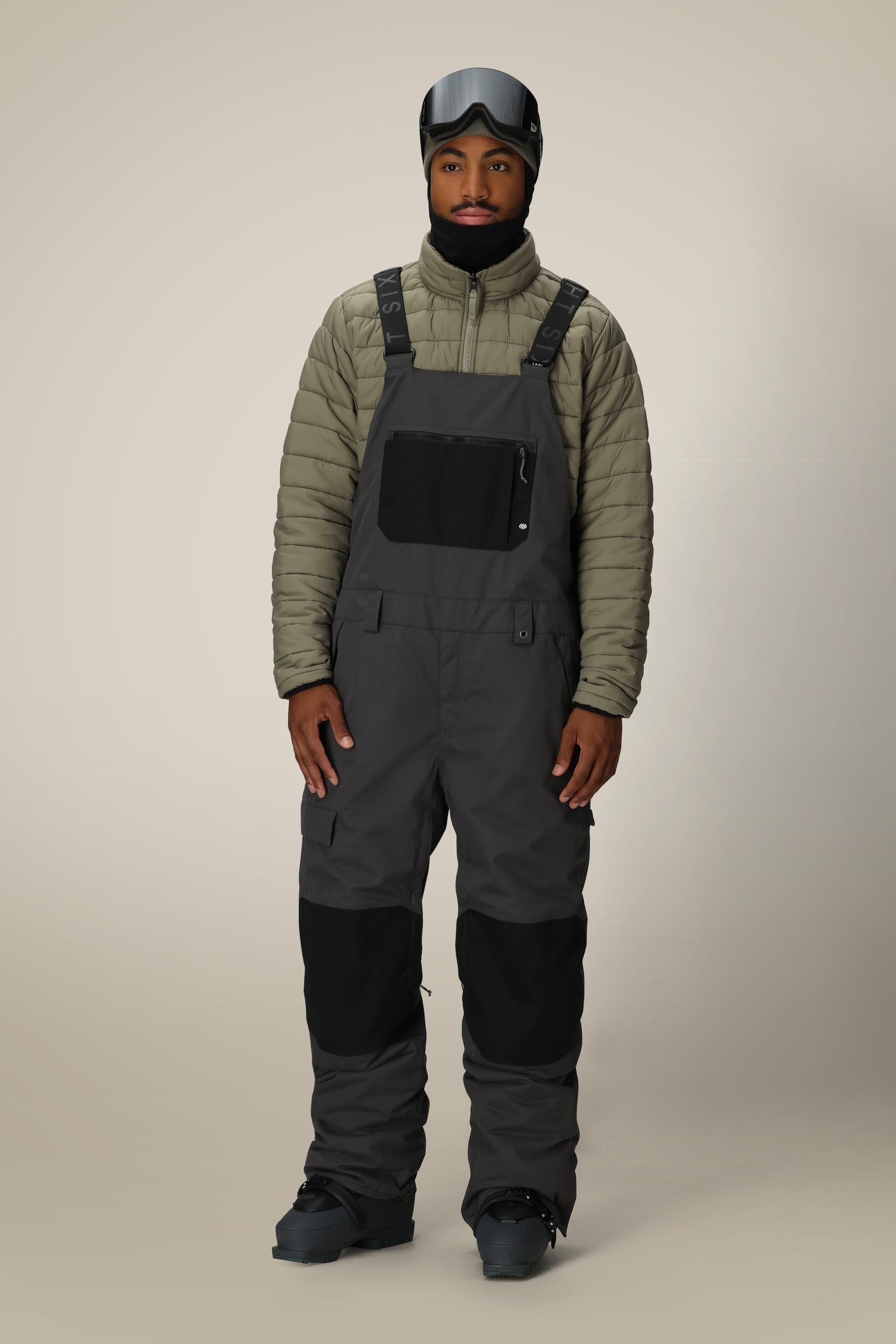 A man stands against a plain beige background wearing the 686 Men's Hot Lap™ Insulated Bib, a gray insulated bib made from polyester nylon spandex with black knee patches, paired with black boots, gloves, and a black ski helmet with goggles.
