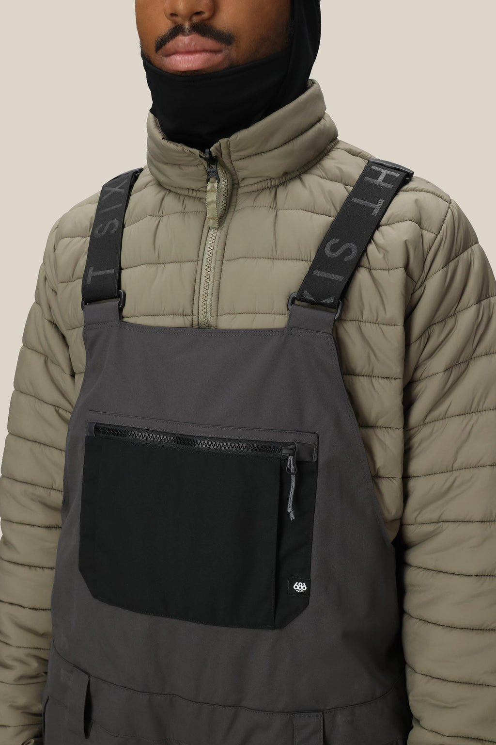 A person wears the 686 Men's Hot Lap™ Insulated Bib by 686, featuring black overalls with a large zippered pocket and logo suspenders, paired with a light olive quilted jacket with a front zipper, against a neutral background.