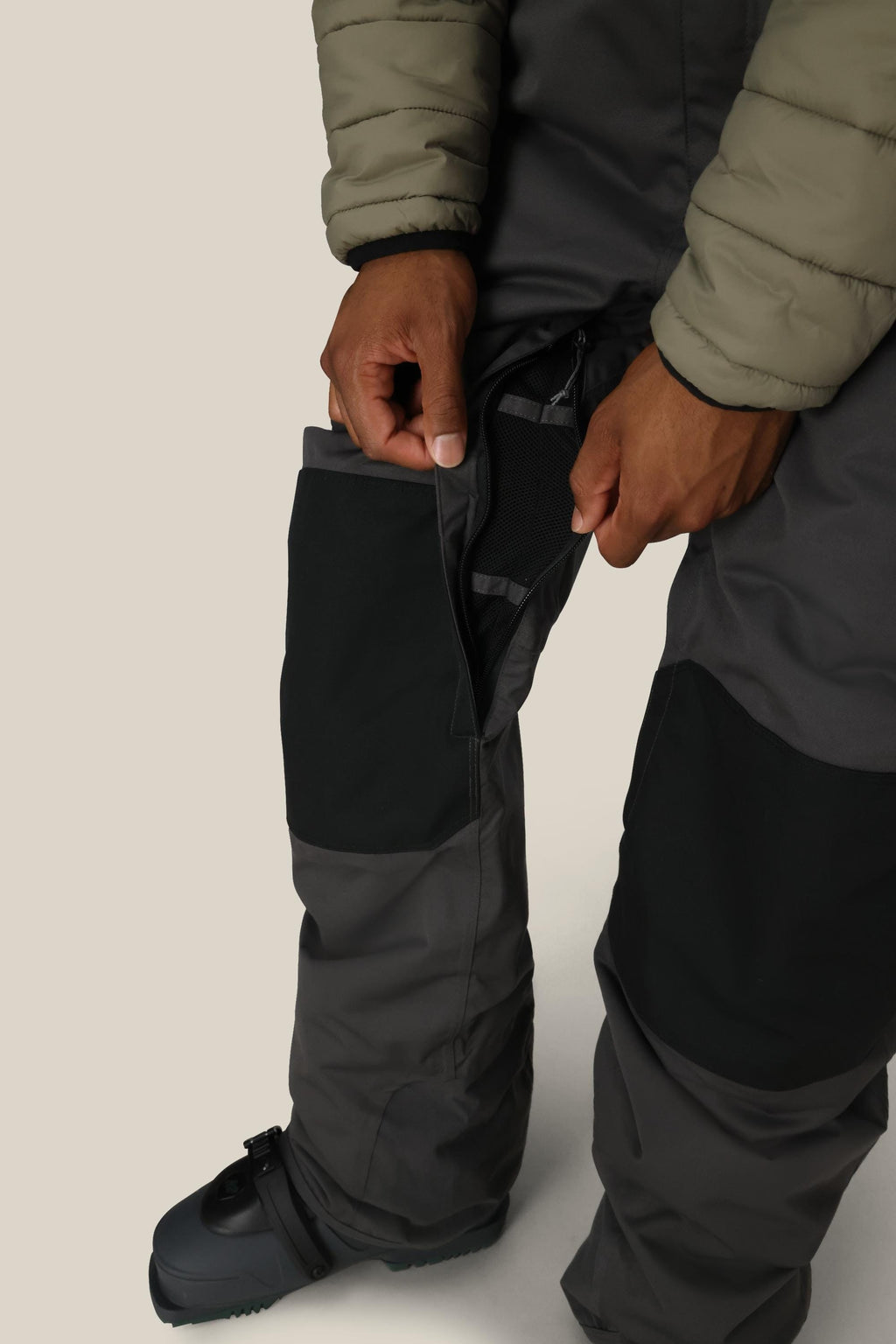 A person wearing 686 Men's Hot Lap™ Insulated Bib in gray and black polyester/nylon/spandex opens a zippered thigh vent, with their hands, legs, and boot partly visible against a plain background. They're also wearing a light green puffer jacket.