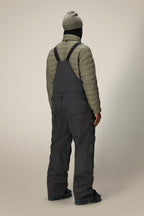 A person stands facing away, wearing a gray beanie, olive-green quilted jacket, and 686 Men's Hot Lap™ Insulated Bib by 686. The waterproof polyester nylon spandex bib stands out against the plain, light-colored background.