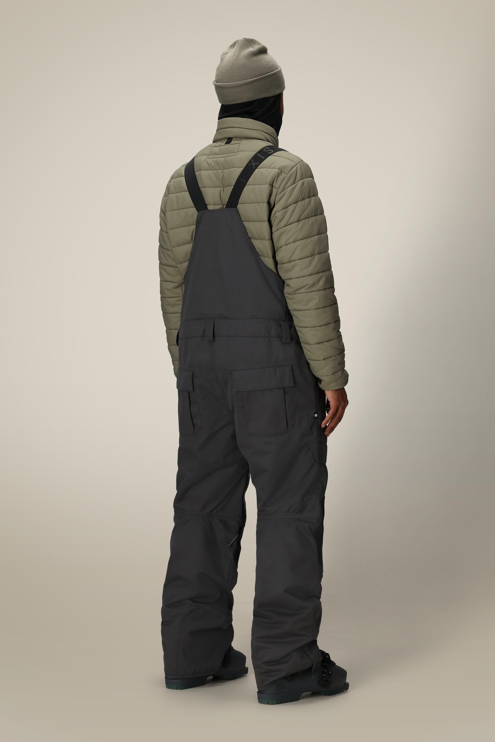 A person stands facing away, wearing a gray beanie, olive-green quilted jacket, and 686 Men's Hot Lap™ Insulated Bib by 686. The waterproof polyester nylon spandex bib stands out against the plain, light-colored background.