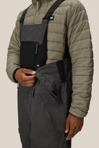 A person in a light olive quilted jacket unfastens the side buttons of the 686 Men's Hot Lap™ Insulated Bib, revealing a black zipper pocket on the chest.