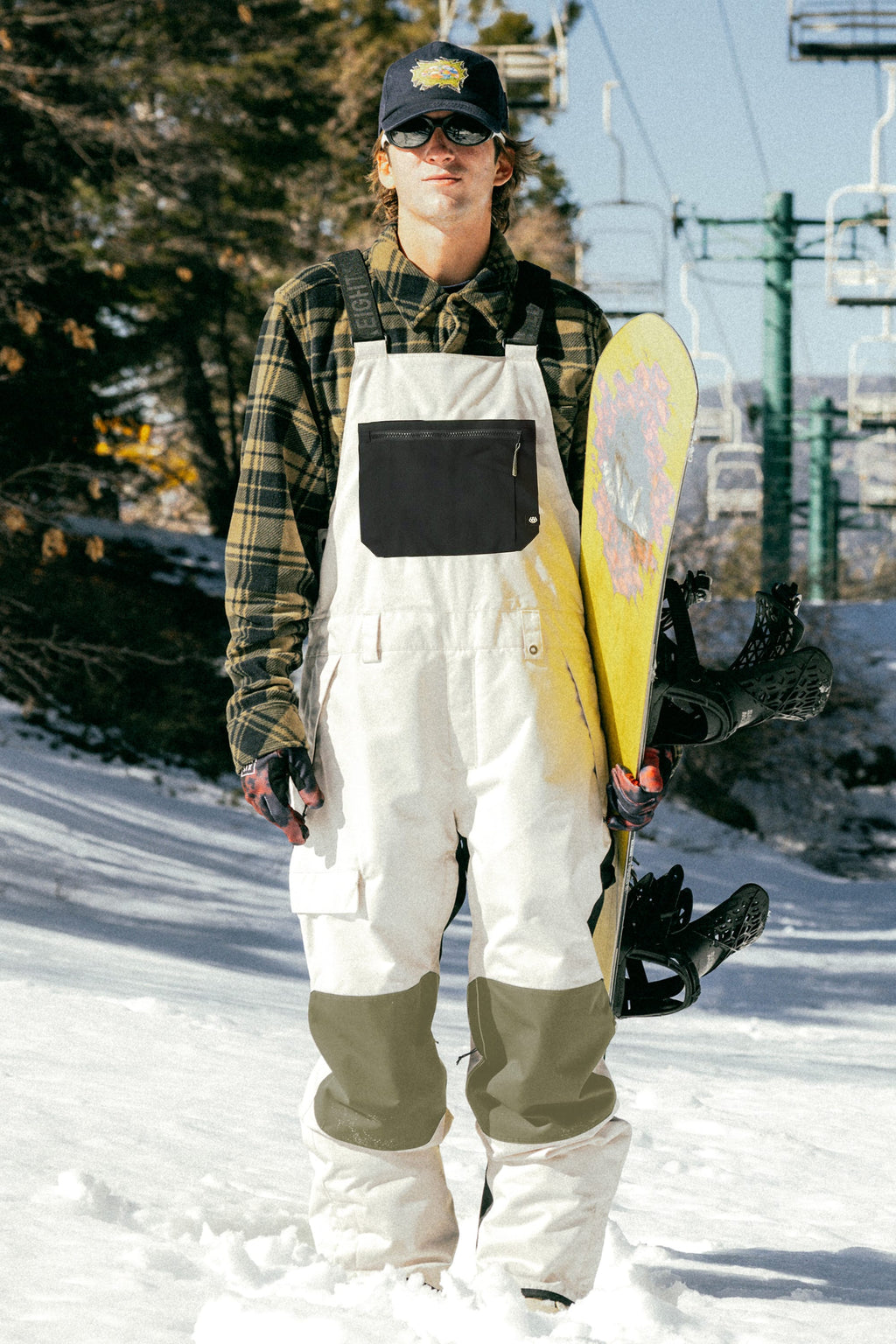 Wearing sunglasses, a cap, and a plaid shirt, a man stands on snowy ground holding a yellow snowboard, dressed in the 686 Men's Hot Lap™ Insulated Bib by 686; ski lift and trees are visible in the background.