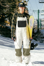 Wearing sunglasses, a cap, and a plaid shirt, a man stands on snowy ground holding a yellow snowboard, dressed in the 686 Men's Hot Lap™ Insulated Bib by 686; ski lift and trees are visible in the background.