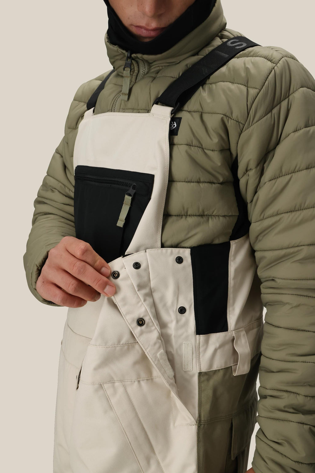A person wearing the 686 Men's Hot Lap™ Insulated Bib by 686 in cream demonstrates snapping shut the side buttons, with only the lower half of their face visible.