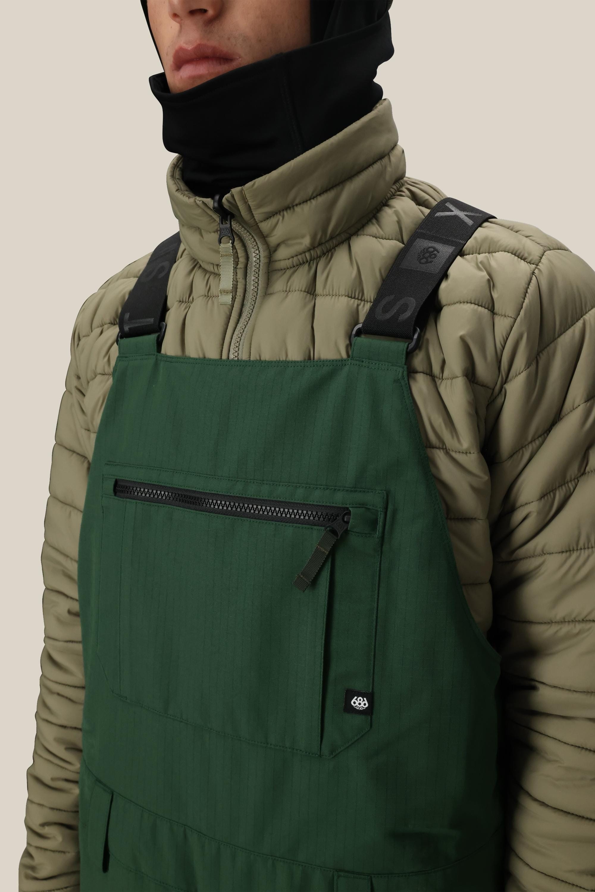 A person wears the 686 Men's Hot Lap™ Insulated Bib by 686, featuring black logo straps, polyester nylon spandex construction, a front zipper pocket, a khaki quilted jacket, and a black neck gaiter covering their neck and lower face.