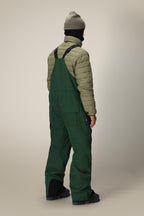 A person stands facing away, wearing a green beanie, a tan quilted jacket, black boots, and dark green 686 Men's Hot Lap™ Insulated Bib overalls with black straps against a neutral background.