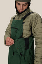 A person wearing the 686 Men's Hot Lap™ Insulated Bib adjusts the buttons on the front pocket flap of the overalls. The background is plain and neutral.
