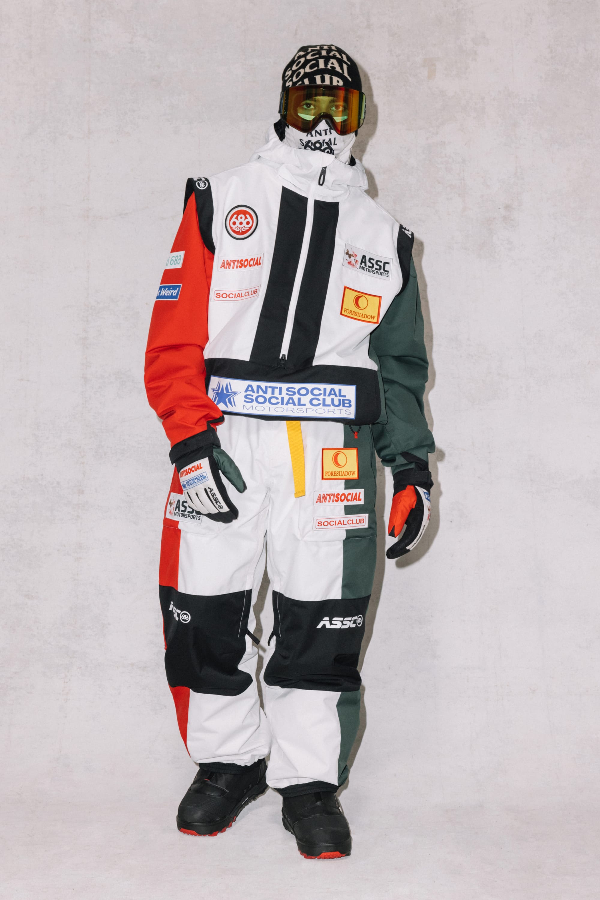 A person wears a colorful snowsuit with red, black, white, and green panels, branded patches, black gloves and boots, and ski goggles. The outfit features 686 Mens ASSC F1 Shell Pant in polyester. The background is plain and light-colored.