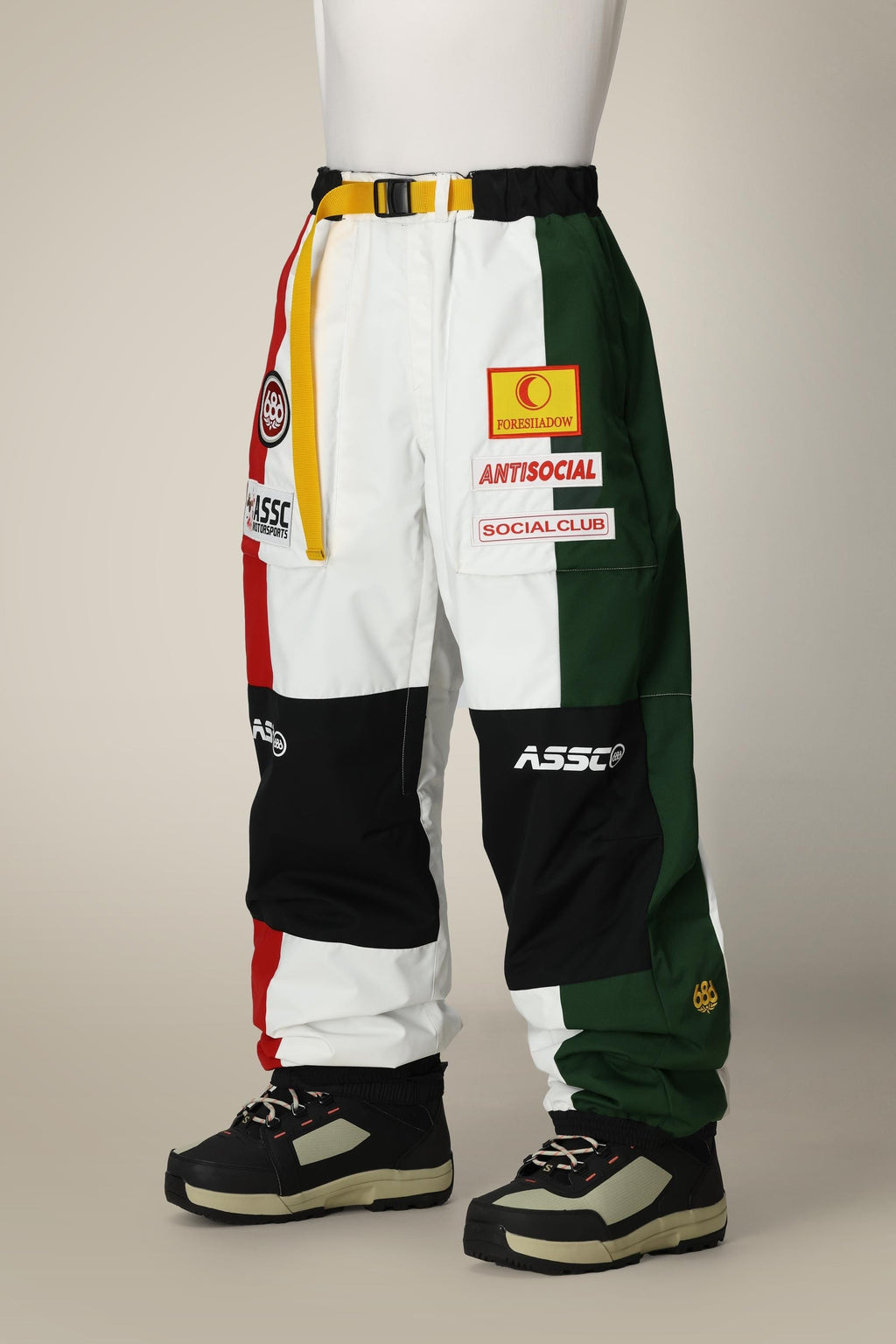 Wearing 686 Mens ASSC F1 Shell Pant by 686, featuring bold red, white, yellow, green, and black color-blocked panels with standout patches and logos, paired with black and beige sneakers. Upper body not shown.