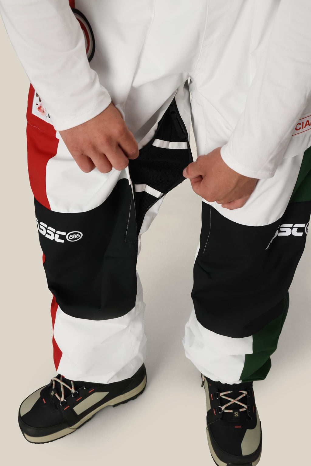 A person wearing 686 Mens ASSC F1 Shell Pant, featuring a red, white, green, and black color-blocked design with zippered vents, unzips the side of one leg. Black and white outdoor shoes complete the look.