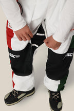A person wearing 686 Mens ASSC F1 Shell Pant, featuring a red, white, green, and black color-blocked design with zippered vents, unzips the side of one leg. Black and white outdoor shoes complete the look.