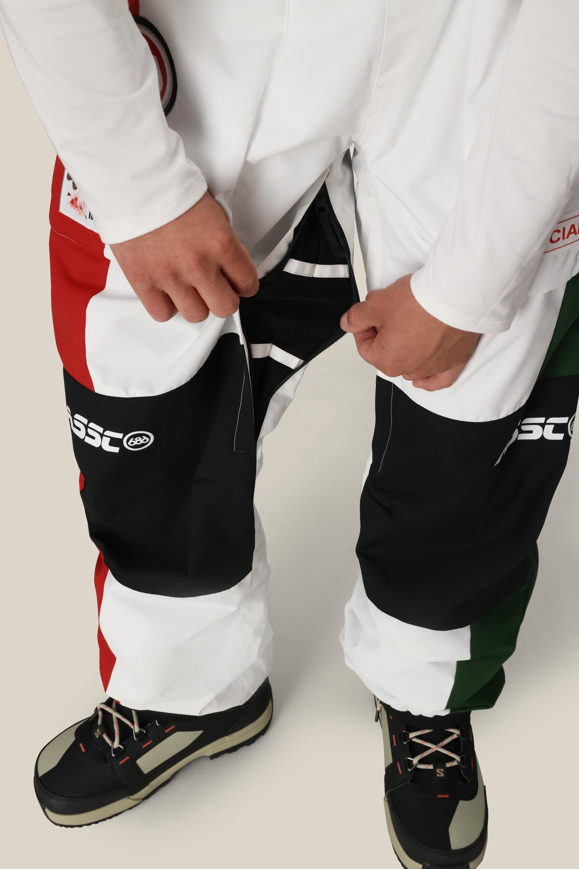 A person wearing 686 Mens ASSC F1 Shell Pant, featuring a red, white, green, and black color-blocked design with zippered vents, unzips the side of one leg. Black and white outdoor shoes complete the look.