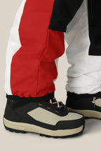A close-up of a person wearing 686 Mens ASSC F1 Shell Pant by 686, featuring red, black, and white color-blocking, paired with beige-black chunky sneakers and standing on a light background.