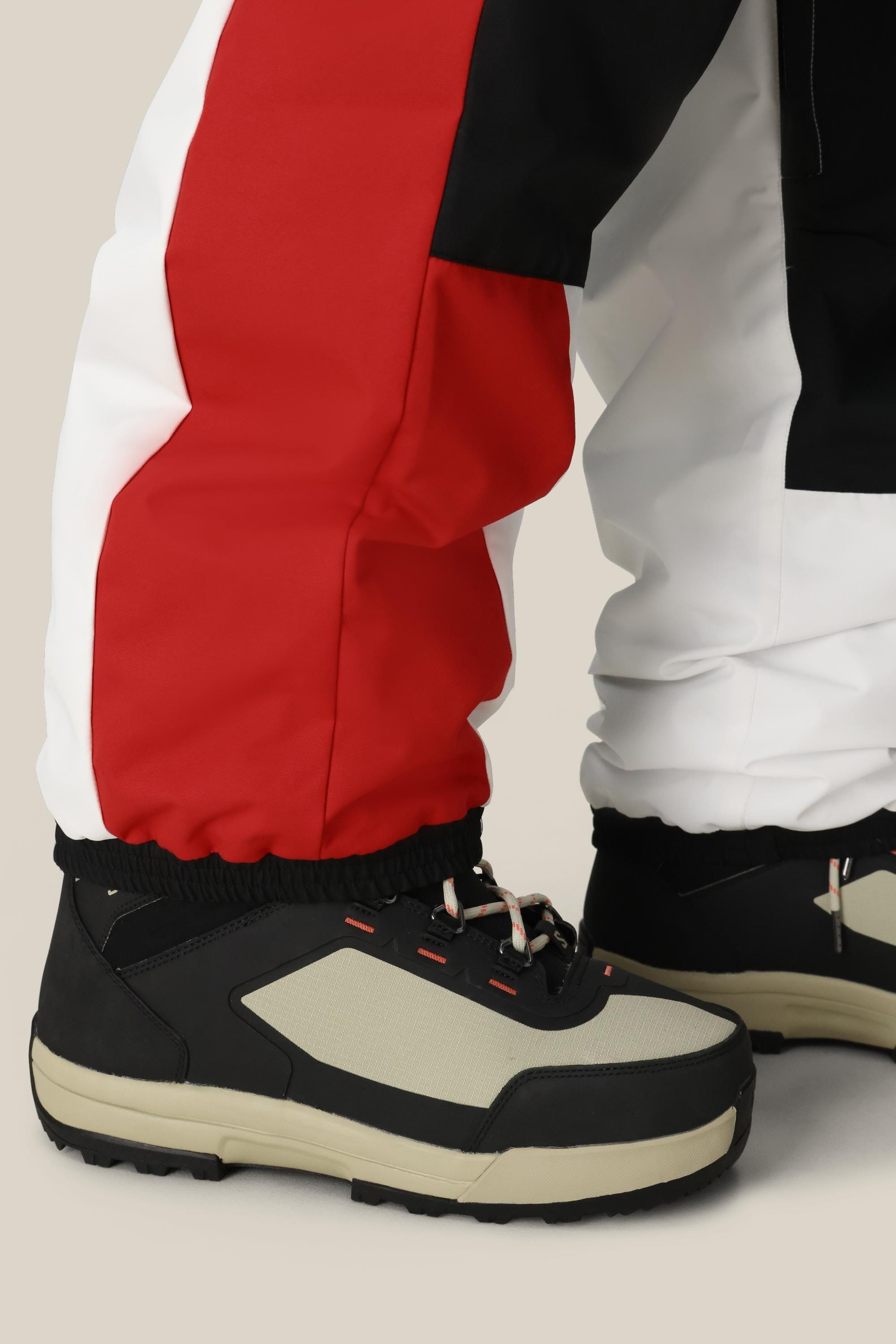 A close-up of a person wearing 686 Mens ASSC F1 Shell Pant by 686, featuring red, black, and white color-blocking, paired with beige-black chunky sneakers and standing on a light background.