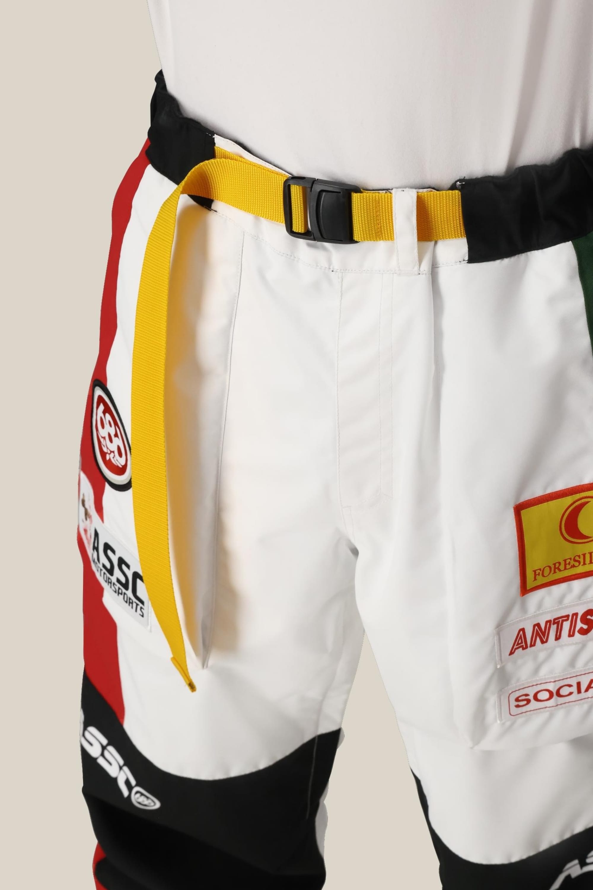 A close-up of a person wearing the 686 Mens ASSC F1 Shell Pant—polyester race pants by 686, in white with red and black accents, colorful sponsor patches, a yellow belt, and a tucked-in white shirt.