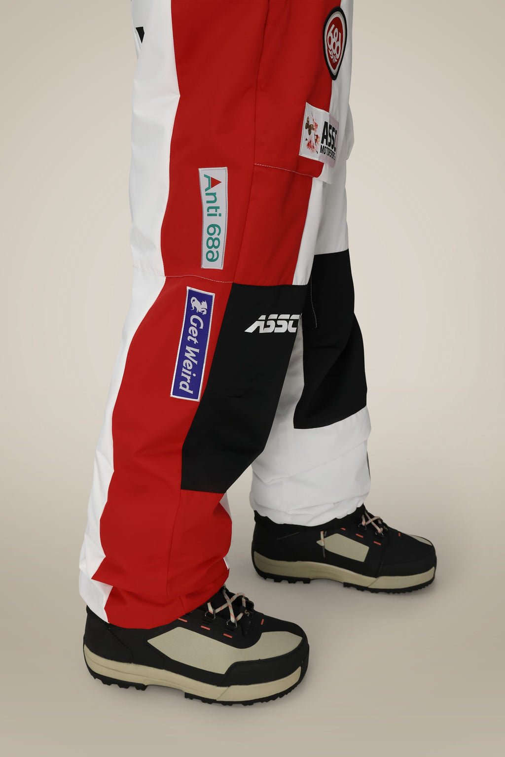 A person wearing 686 Mens ASSC F1 Shell Pant by 686, featuring a red, white, and black color-block design with patches and logos, paired with black and beige boots on a neutral beige background.