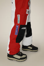 A person wearing 686 Mens ASSC F1 Shell Pant by 686, featuring a red, white, and black color-block design with patches and logos, paired with black and beige boots on a neutral beige background.