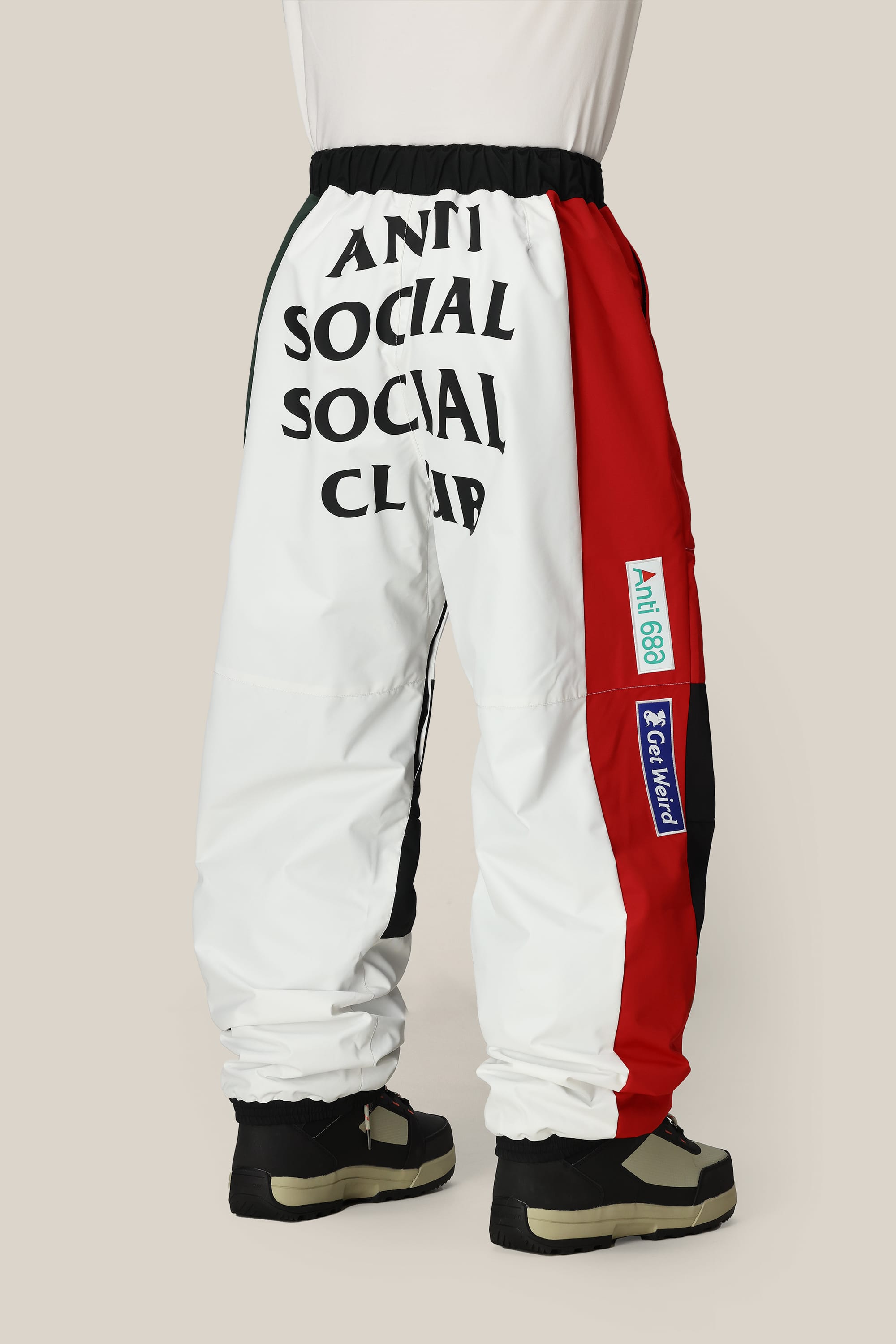 A person wears the 686 Mens ASSC F1 Shell Pant by 686, featuring a white, black, and red color-block design with “ANTI SOCIAL SOCIAL CLUB” on the back and blue, green, and white leg patches; styled with black and beige sneakers.