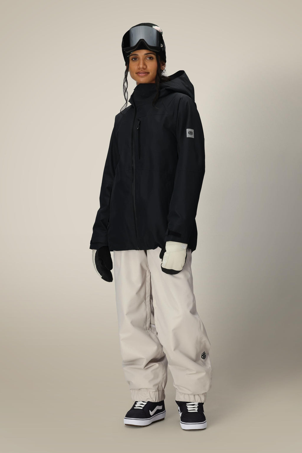 A person wearing the 686 Women's Hydra™ Thermagraph® Jacket, white snow pants, black and white gloves, black and white sneakers, and ski goggles stands against a plain background.