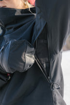 A person outdoors unzips the underarm vent of the black 686 Women's Hydra™ Thermagraph® Jacket, revealing mesh lining. They are wearing a black padded glove, and a hint of a red hat is visible.