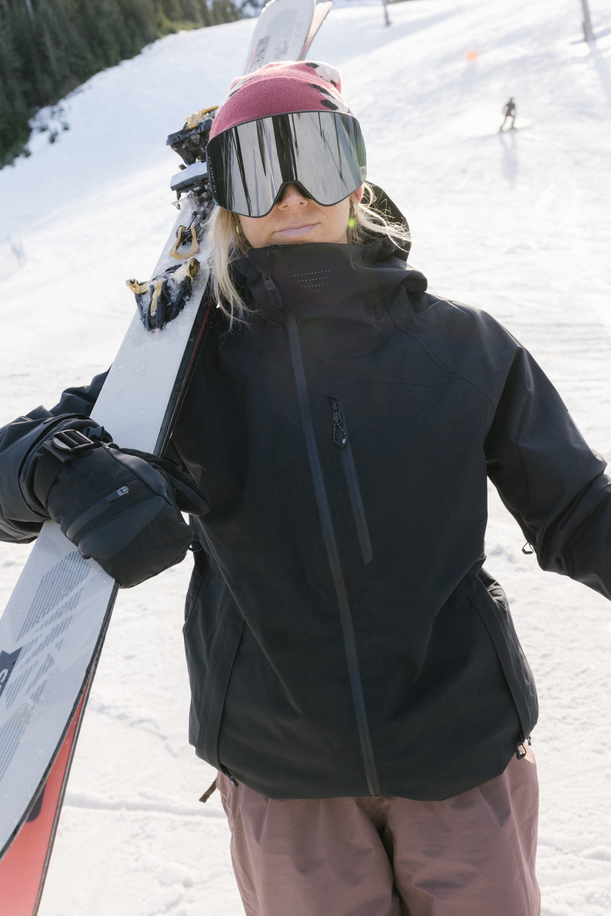 A skier in a 686 Women's Hydra™ Thermagraph® Jacket, pink beanie, and reflective goggles stands on a snowy slope with skis on her shoulder. Pine trees and another skier appear in the background.