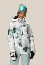 A woman wearing the 686 Women's Hydra™ Thermagraph® Jacket, gray snow pants, ski gloves, a beanie, and ski goggles on her head stands against a plain beige background, smiling slightly.