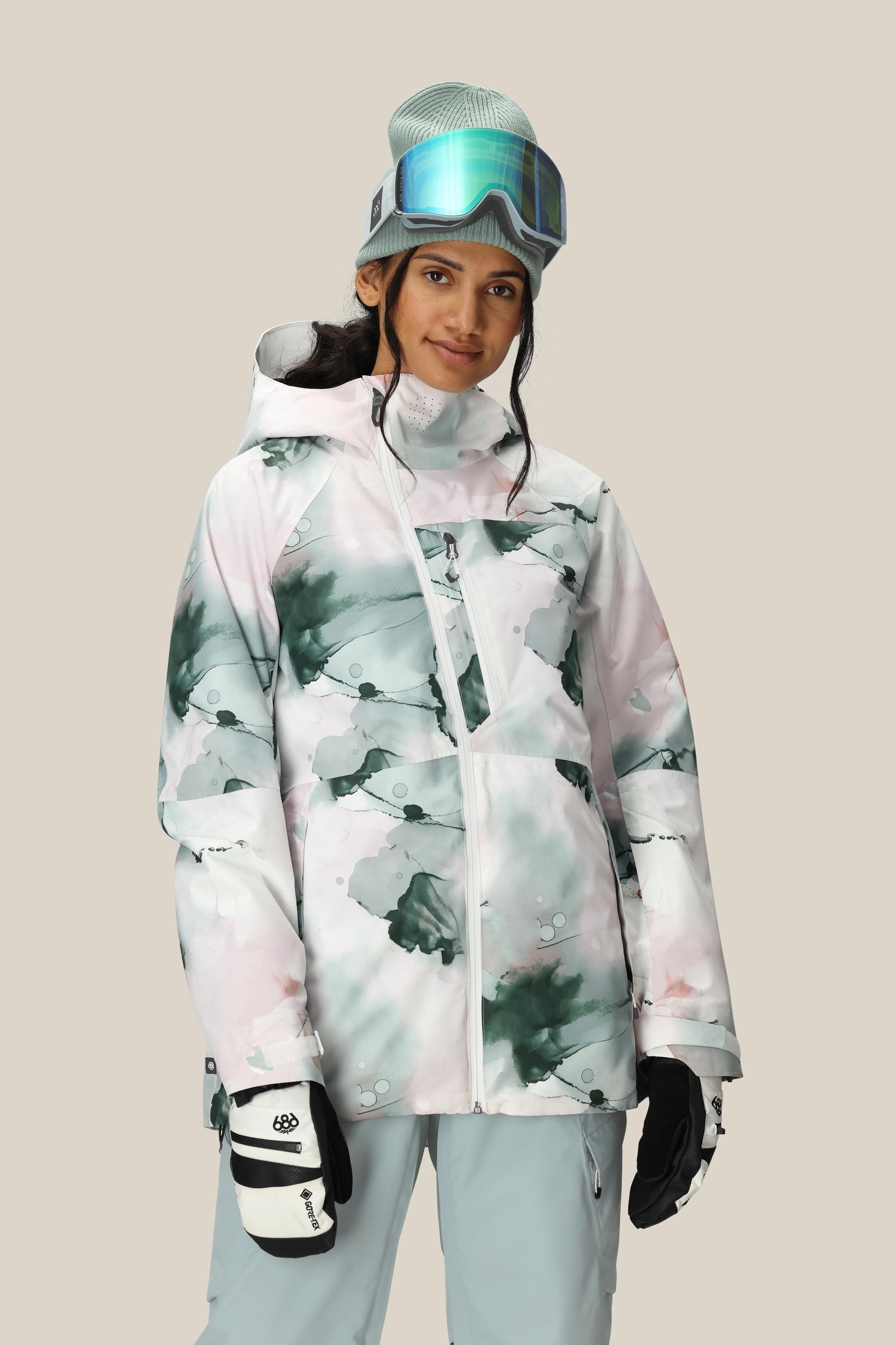 A woman wearing the 686 Women's Hydra™ Thermagraph® Jacket, gray snow pants, ski gloves, a beanie, and ski goggles on her head stands against a plain beige background, smiling slightly.