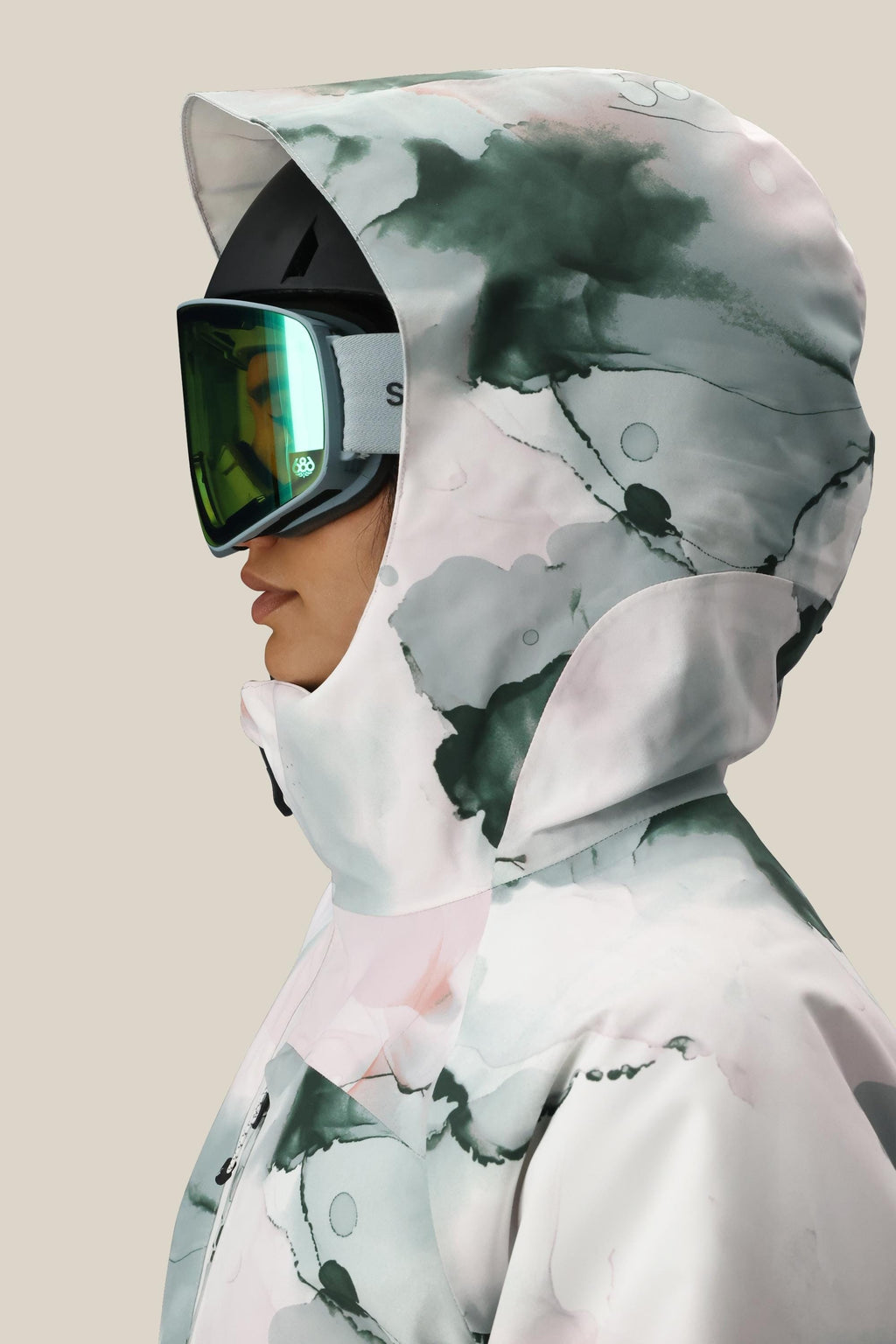 A person in profile wears the 686 Women's Hydra™ Thermagraph® Jacket, featuring a watercolor pattern, paired with reflective ski goggles against a plain beige background.