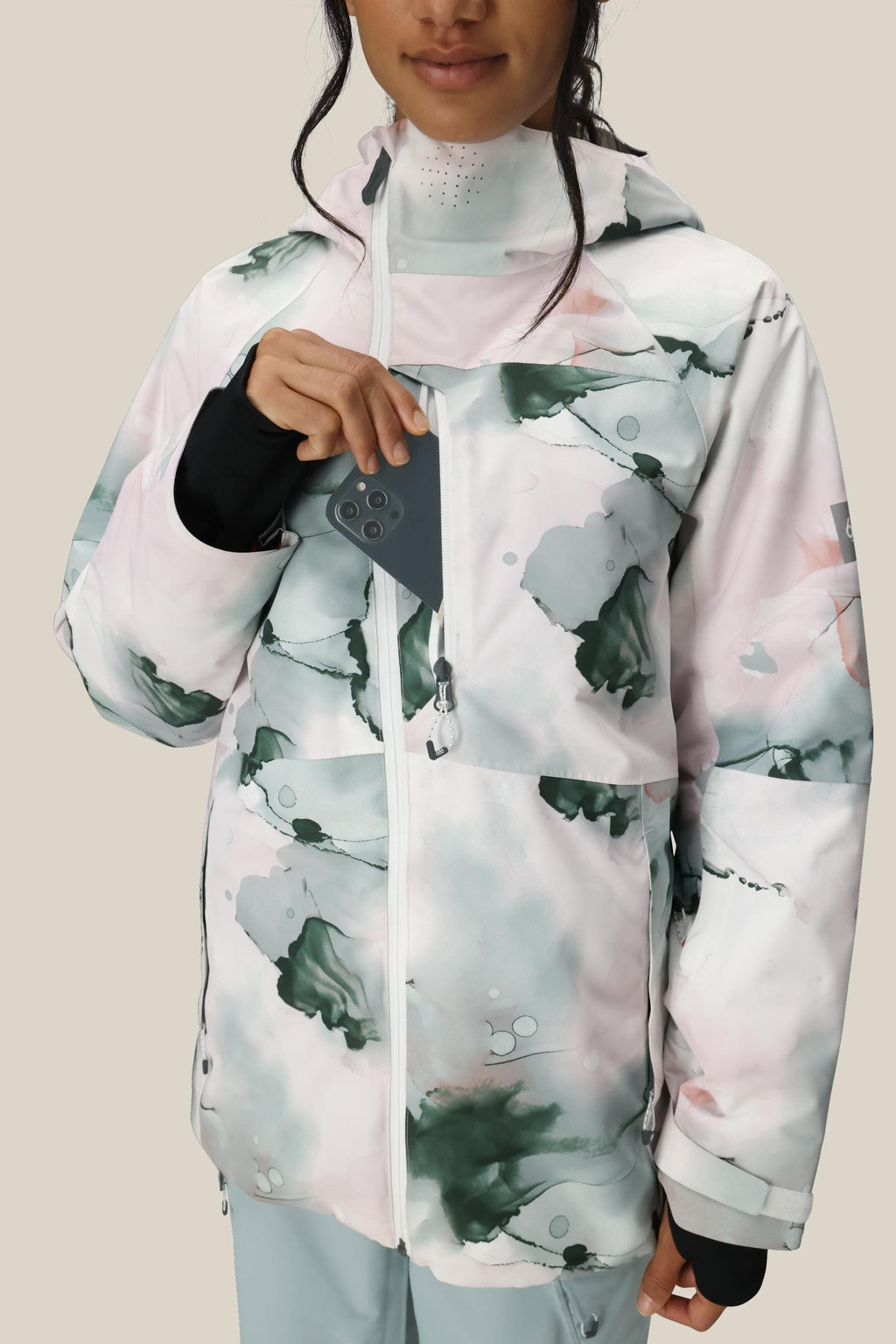 A woman wearing the 686 Women's Hydra™ Thermagraph® Jacket in a white and pastel watercolor pattern puts her smartphone into the chest pocket. The jacket features a hood, black wrist gaiters, and is set against a plain beige background.