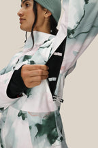 A woman in a green beanie unzips an underarm vent on the 686 Women's Hydra™ Thermagraph® Jacket, revealing mesh lining. The background is plain and neutral.