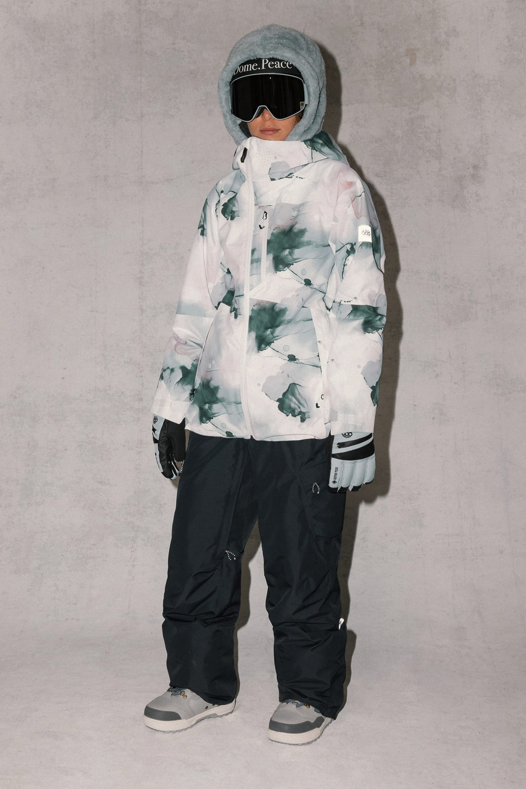 A child stands against a light gray background wearing the 686 Women's Hydra™ Thermagraph® Jacket in a gray, white, and green abstract pattern by 686, paired with black snow pants, gray gloves and boots, and ski goggles.