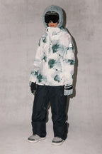 A child stands against a light gray background wearing the 686 Women's Hydra™ Thermagraph® Jacket in a gray, white, and green abstract pattern by 686, paired with black snow pants, gray gloves and boots, and ski goggles.