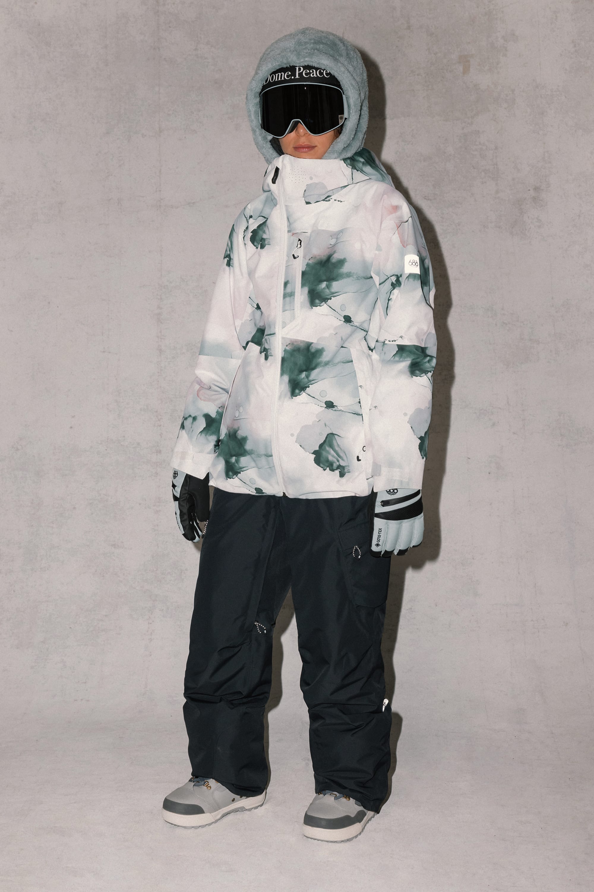 A child stands against a light gray background wearing the 686 Women's Hydra™ Thermagraph® Jacket in a gray, white, and green abstract pattern by 686, paired with black snow pants, gray gloves and boots, and ski goggles.