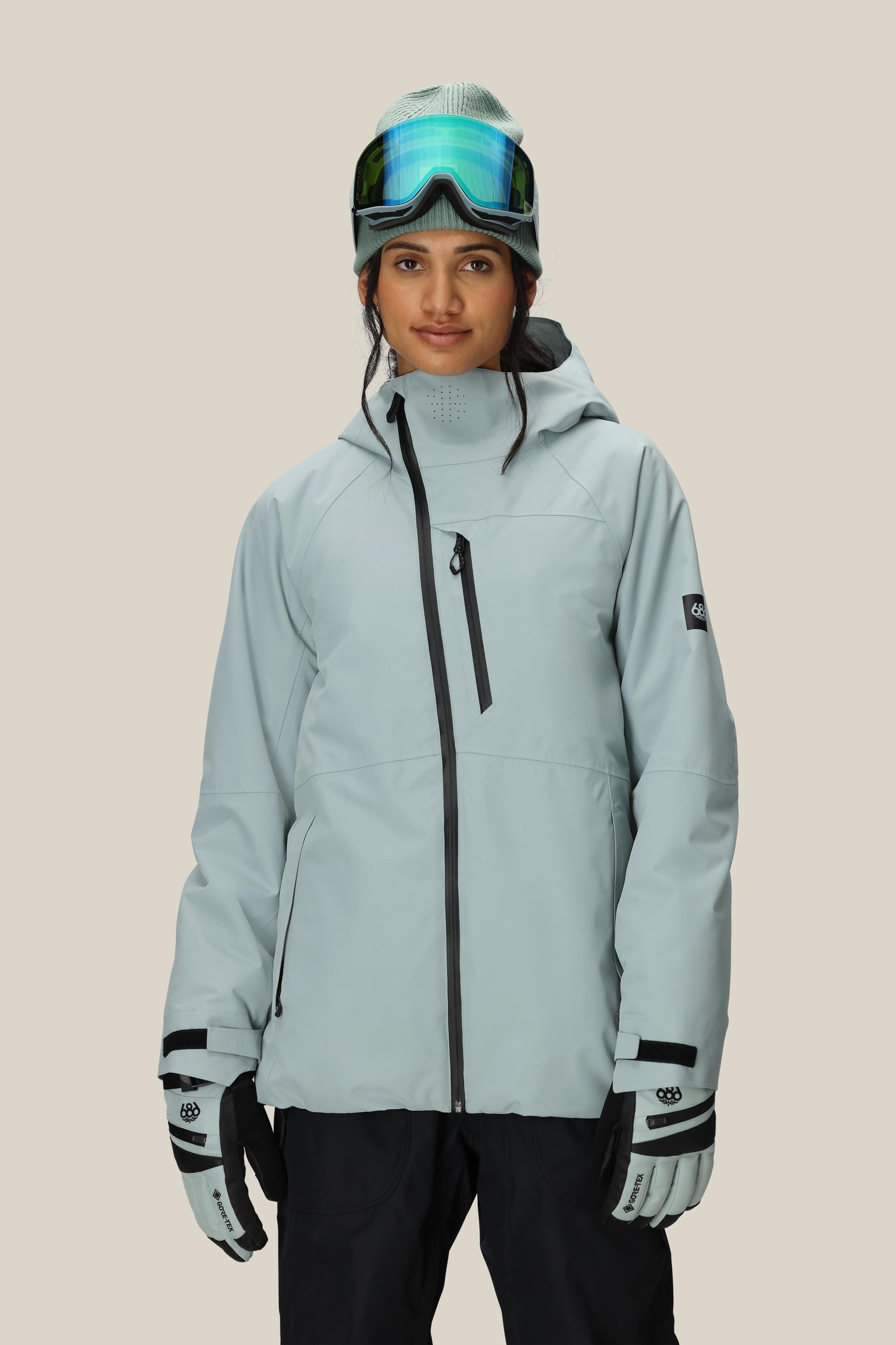 A person wearing the 686 Women's Hydra™ Thermagraph® Jacket in light gray, black pants, gloves, and ski goggles on their head stands against a plain beige background.