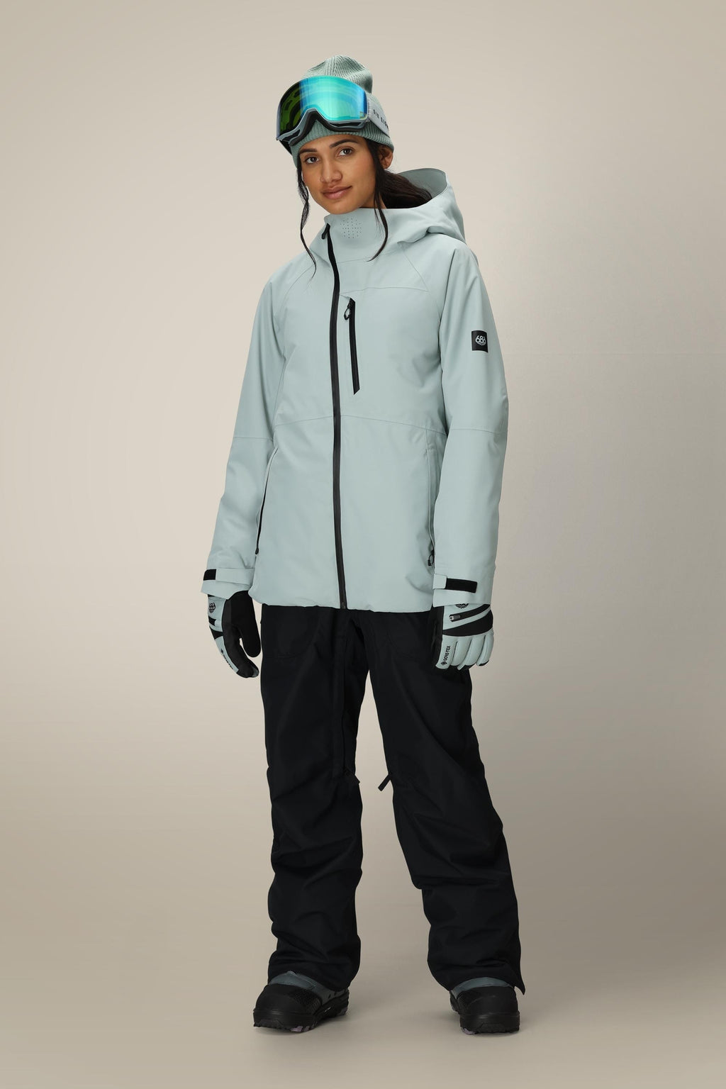 A woman stands facing forward against a plain, light background, wearing the 686 Women's Hydra™ Thermagraph® Jacket in light gray with a hood, black snow pants, ski gloves, goggles, and a beanie.