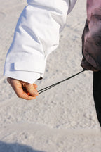 A person wearing the 686 Women's Hydra™ Thermagraph® Jacket adjusts a black drawstring while standing on snow or ice. Only their hand, arm, and part of the 686 waterproof jacket are visible.