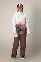 A person stands facing forward against a plain background, wearing the 686 Women's Hydra™ Thermagraph® Jacket with a black abstract design in white and pink, brown snow pants, white gloves, black sneakers, and ski goggles.