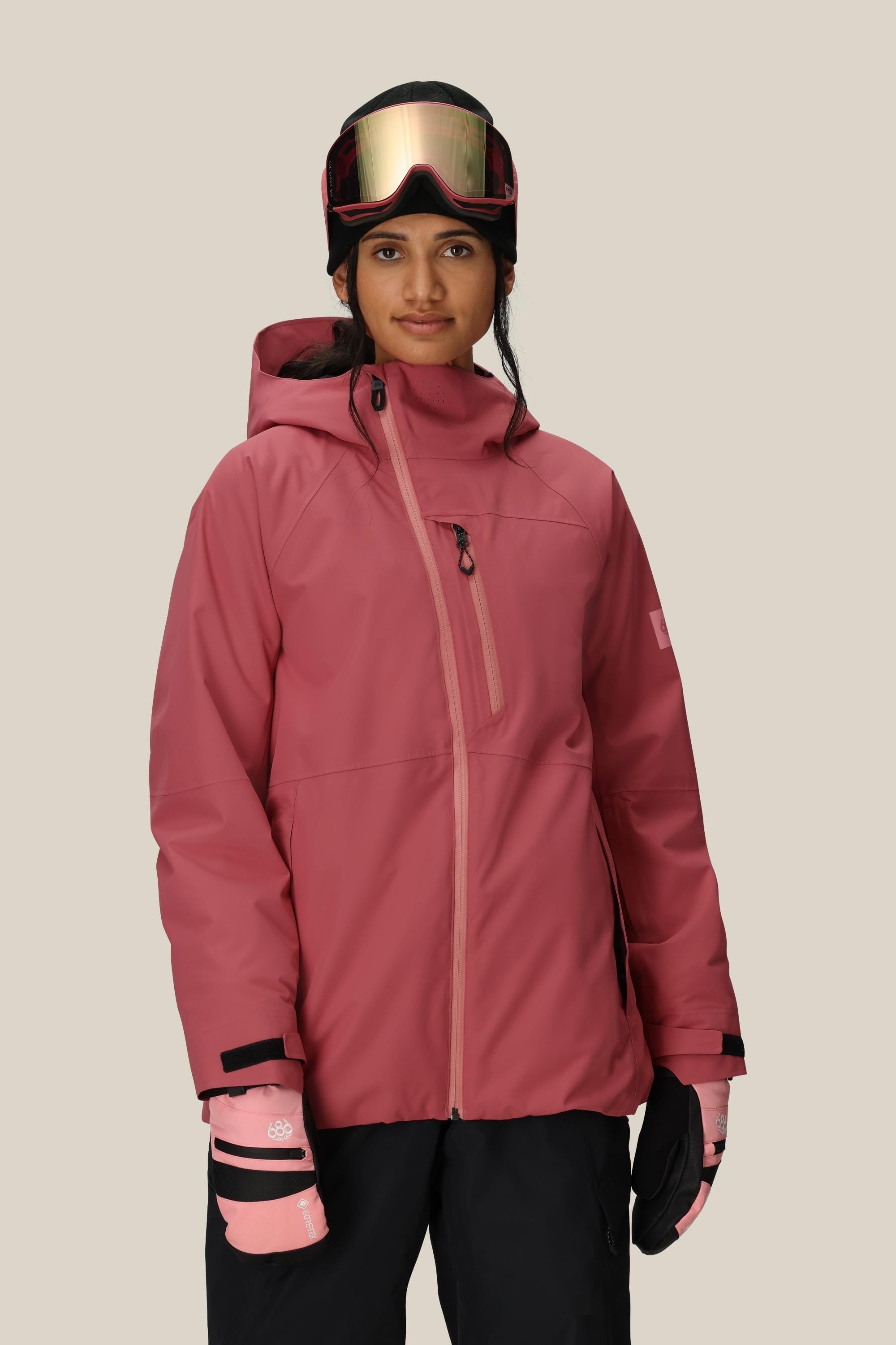 A person wears a rose pink 686 Women's Hydra™ Thermagraph® Jacket, matching gloves, black snow pants, and a ski helmet with goggles, standing against a plain beige background.