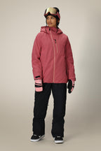 A person in winter attire wears the 686 Women's Hydra™ Thermagraph® Jacket in pink with hood, black snow pants, pink gloves, black shoes, and ski goggles on their head, standing against a plain light background.