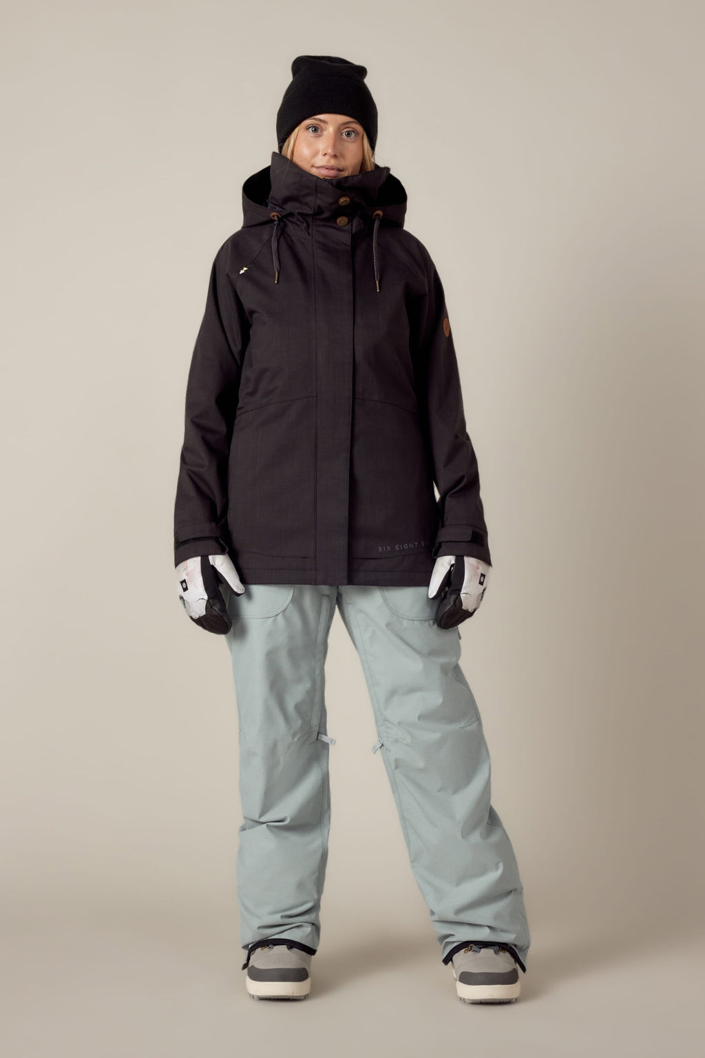 A person stands indoors against a plain beige background, wearing the 686 Women's SMARTY® 3-In-1 Spellbound Jacket in black, light blue snow pants, a black beanie, white gloves, and snow boots, looking directly at the camera.