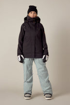 A person stands indoors against a plain beige background, wearing the 686 Women's SMARTY® 3-In-1 Spellbound Jacket in black, light blue snow pants, a black beanie, white gloves, and snow boots, looking directly at the camera.