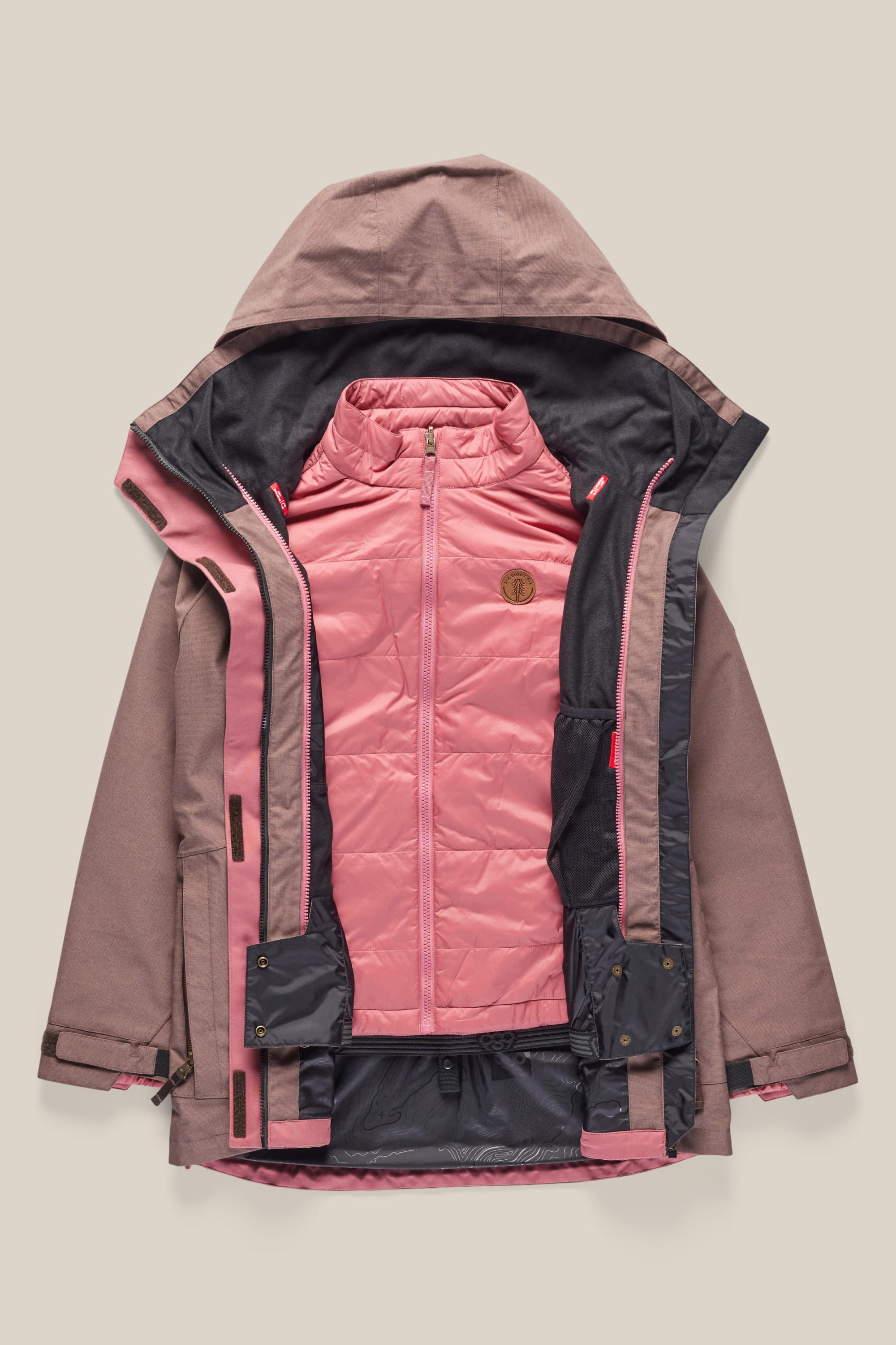 The 686 Women's SMARTY® 3-In-1 Spellbound Jacket by 686 is shown unzipped, revealing its quilted light pink inner layer and stand-up collar, with a brown logo patch on the chest, all set against a beige background.