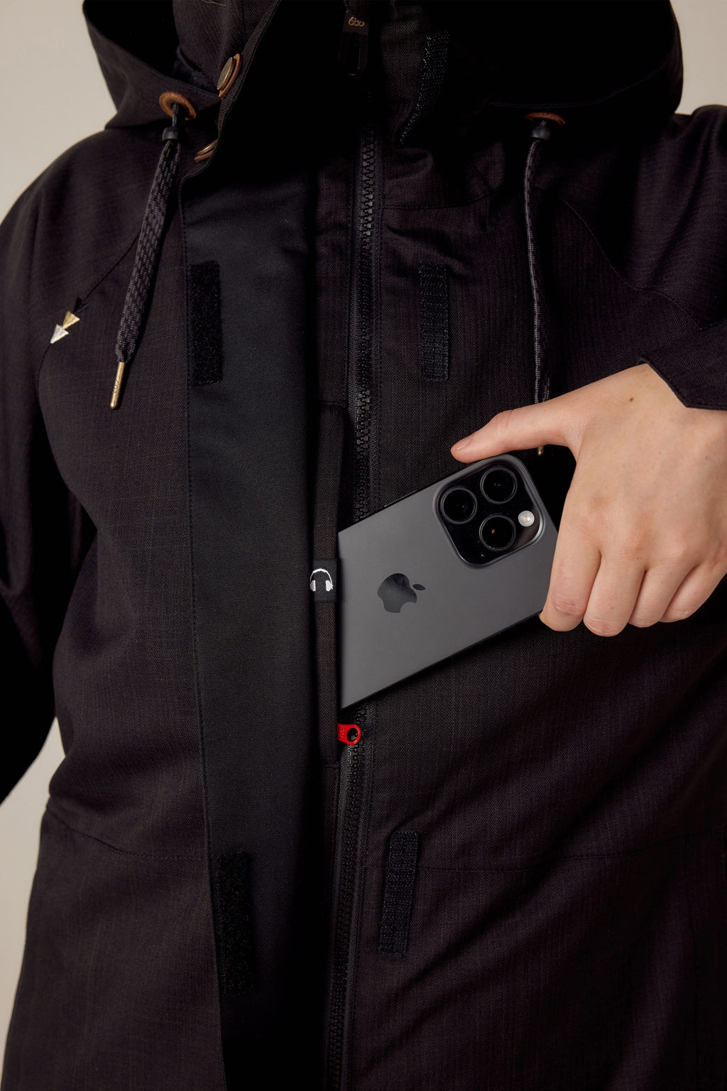 A woman in a black 686 Women's SMARTY® 3-In-1 Spellbound Jacket slips a black iPhone with three cameras into an inner chest pocket, her jacket unzipped halfway to show the pocket.