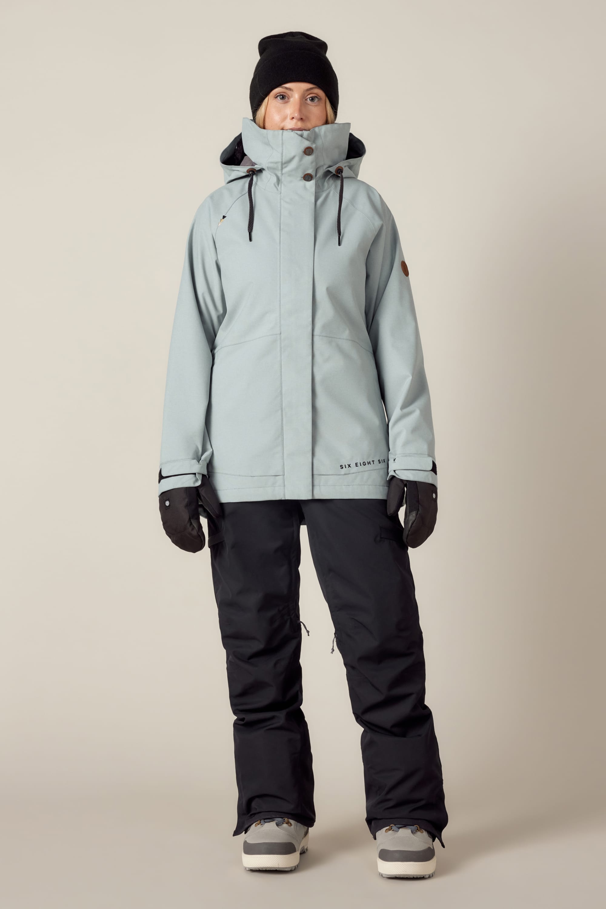 A person stands facing forward, wearing the 686 Women's SMARTY® 3-In-1 Spellbound Jacket by 686, black snow pants, black gloves, a black beanie, and gray winter boots against a plain light background.