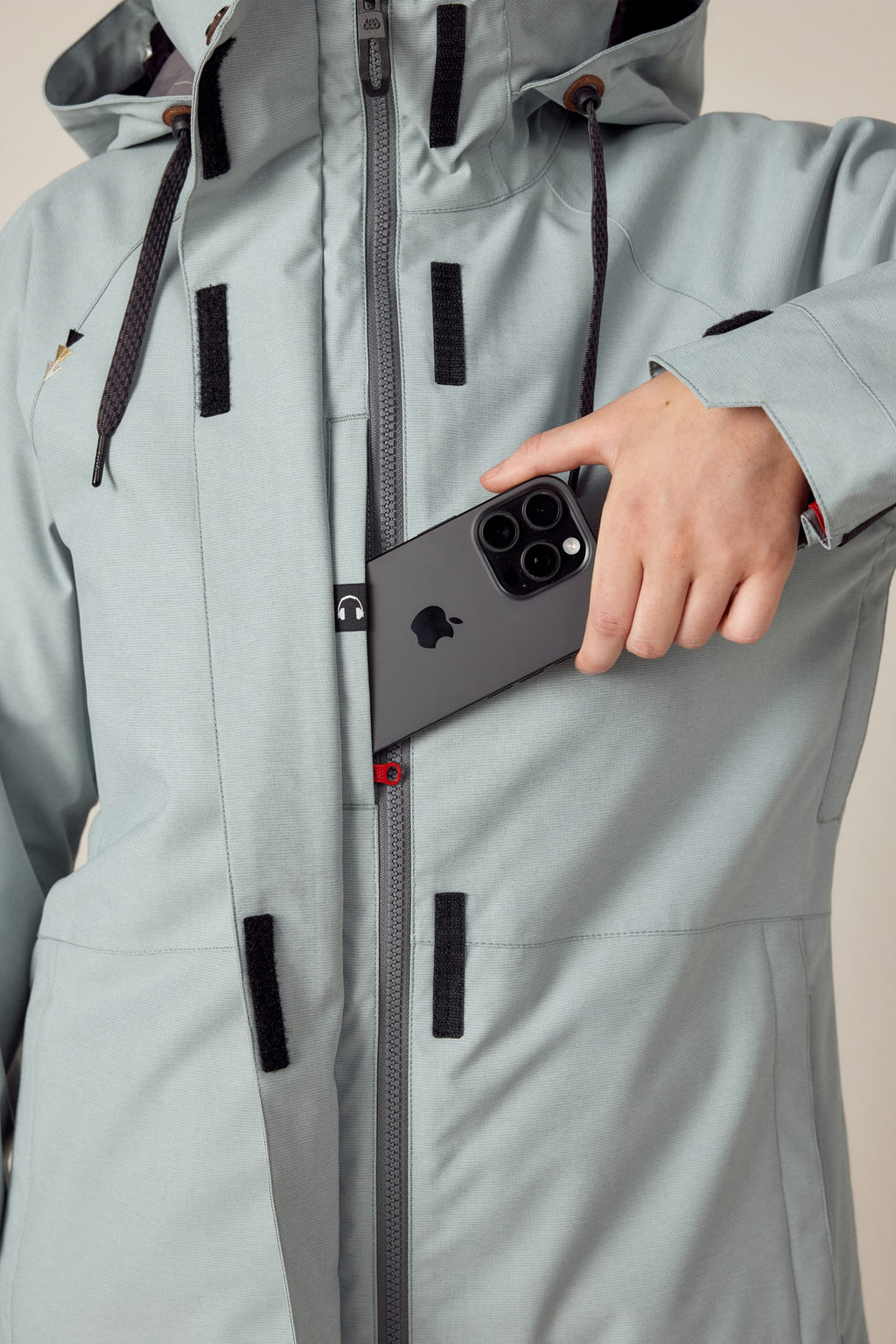 A woman in a light gray 686 Women's SMARTY® 3-In-1 Spellbound Jacket with black zippers, velcro patches, and hood places a triple-camera black smartphone into the jacket’s chest pocket.
