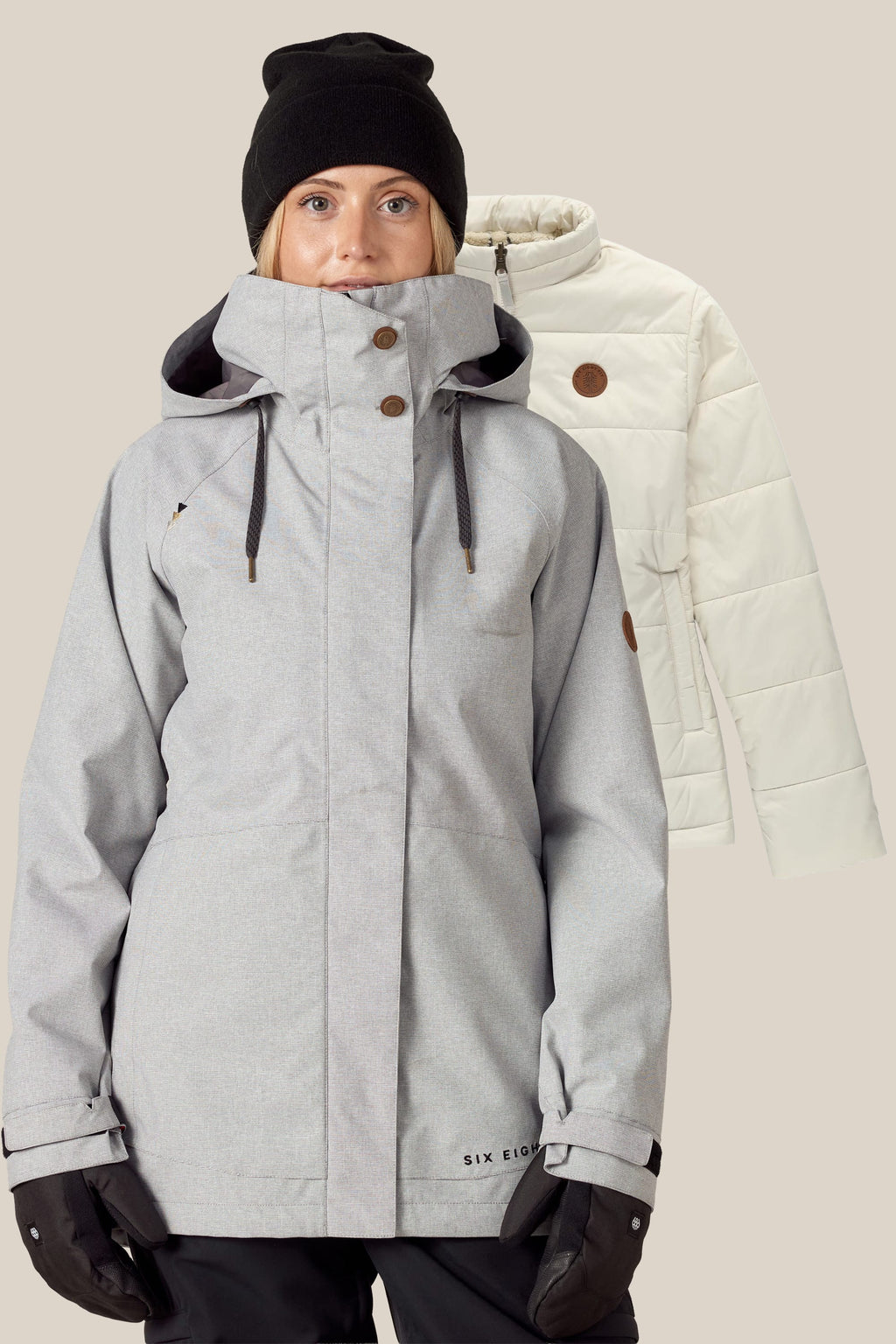 A woman in a gray winter jacket, black gloves, and a black beanie stands in front of the 686 Women's SMARTY® 3-In-1 Spellbound Jacket by 686 in light beige, displayed against a plain background.