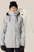 A woman in a gray winter jacket, black gloves, and a black beanie stands in front of the 686 Women's SMARTY® 3-In-1 Spellbound Jacket by 686 in light beige, displayed against a plain background.