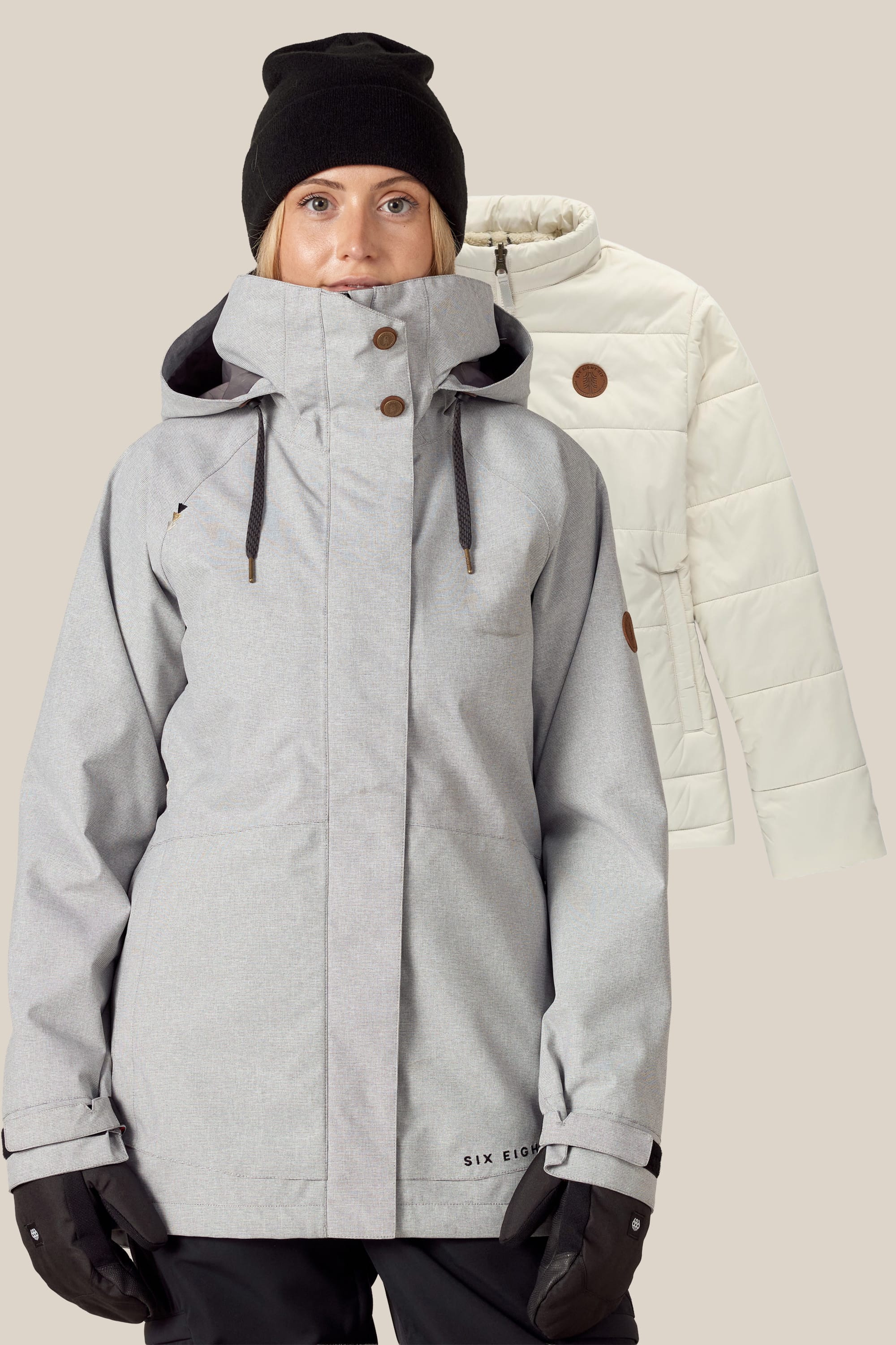 A woman in a gray winter jacket, black gloves, and a black beanie stands in front of the 686 Women's SMARTY® 3-In-1 Spellbound Jacket by 686 in light beige, displayed against a plain background.