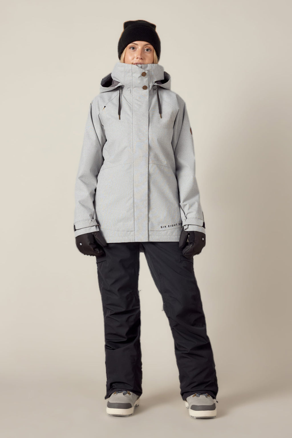 A person faces forward in a 686 Women's SMARTY® 3-In-1 Spellbound Jacket, black gloves, black pants, winter boots, and a black beanie against a plain light background.